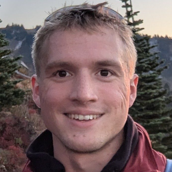 Joel smiling outdoors with mountains and evergreen trees behind him