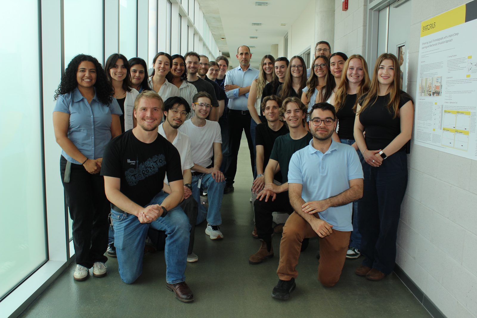 Joel kneeling in the front row with the research team in a McMaster hallway, beside a Sartorius research poster